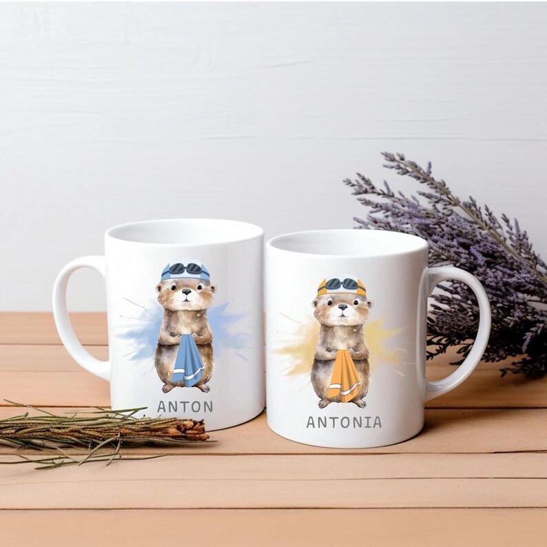 Pode incluir: Duas canecas de cer&acirc;mica branca com ilustra&ccedil;&otilde;es de lontras. Cada lontra usa &oacute;culos de nata&ccedil;&atilde;o e segura uma toalha. Uma caneca diz "ANTON" e a outra "ANTONIA". As lontras est&atilde;o sobre um fundo de aquarela azul e amarelo.