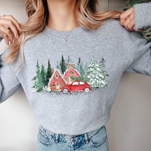 May include: A heather gray crewneck sweatshirt featuring a watercolor-style graphic of two red houses nestled among snow-covered evergreen trees, with a red vintage-style car carrying a Christmas tree on its roof parked in the foreground.