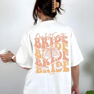 May include: White t-shirt with a retro design featuring the text "One last Spritz" above the word "Bride" repeated multiple times. The design includes two cocktail glasses with orange slices. The name "Kathi" is printed at the bottom.