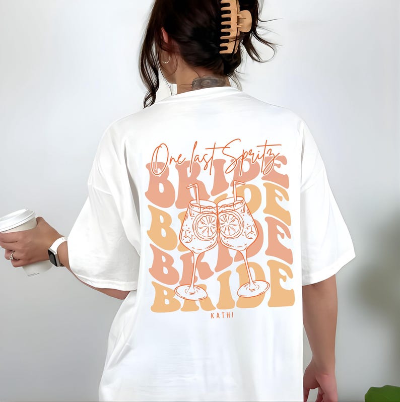 May include: White t-shirt with a retro design featuring the text "One last Spritz" above the word "Bride" repeated multiple times. The design includes two cocktail glasses with orange slices. The name "Kathi" is printed at the bottom.