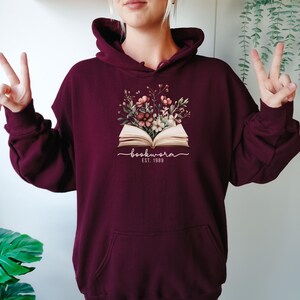May include: Burgundy coloured hoodie featuring a floral design above an open book. The graphic includes the word "bookworm" in cursive and the text "EST. 1989" in white. The hoodie has a front pocket and a drawstring hood.