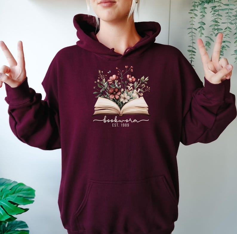 May include: Burgundy coloured hoodie featuring a floral design above an open book. The graphic includes the word "bookworm" in cursive and the text "EST. 1989" in white. The hoodie has a front pocket and a drawstring hood.