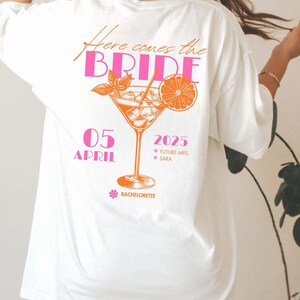 May include: White t-shirt with a pink and orange graphic design. The design includes the text "Here comes the BRIDE" and a cocktail illustration with the date "05 APRIL 2025". The word "Bachelorette" is also included in the design.