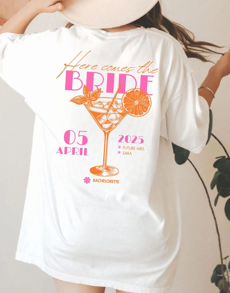 May include: White t-shirt with a pink and orange graphic design. The design includes the text "Here comes the BRIDE" and a cocktail illustration with the date "05 APRIL 2025". The word "Bachelorette" is also included in the design.