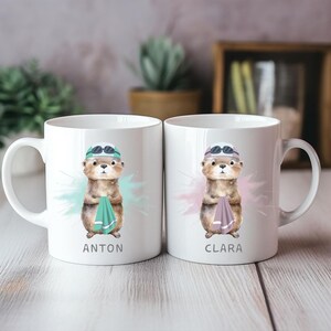 Pode incluir: Duas canecas de cer&acirc;mica branca com uma lontra de desenho animado usando &oacute;culos de nata&ccedil;&atilde;o e touca, segurando uma toalha. Uma caneca diz "ANTON" e a outra "CLARA". Cada caneca tem um design de respingos coloridos.