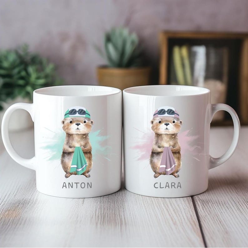 Pode incluir: Duas canecas de cer&acirc;mica branca com uma lontra de desenho animado usando &oacute;culos de nata&ccedil;&atilde;o e touca, segurando uma toalha. Uma caneca diz "ANTON" e a outra "CLARA". Cada caneca tem um design de respingos coloridos.