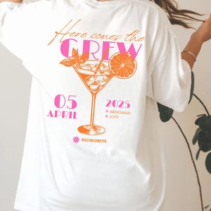 May include: White t-shirt with a cocktail graphic in orange and pink. The text "Here comes the CREW" is above the cocktail. Below the cocktail are the words "05 APRIL 2025" and "Bachelorette".