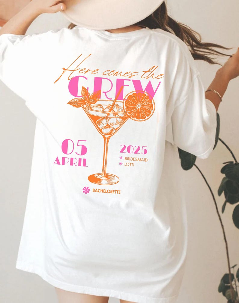 May include: White t-shirt with a cocktail graphic in orange and pink. The text "Here comes the CREW" is above the cocktail. Below the cocktail are the words "05 APRIL 2025" and "Bachelorette".