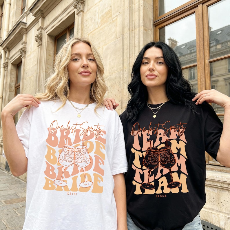 May include: Two women wearing matching t-shirts. One shirt is white with the text "One last Spritz Bride" and a cocktail graphic. The other shirt is black with "One last Spritz Team" and a similar cocktail graphic.