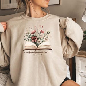 May include: Beige sweatshirt with a graphic of an open book and a floral design. The graphic includes the word "bookworm" and the year "EST. 1989". The sweatshirt is a neutral colour and has long sleeves.