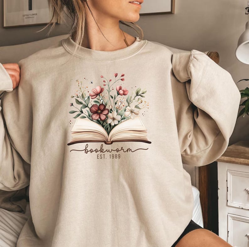May include: Beige sweatshirt with a graphic of an open book and a floral design. The graphic includes the word "bookworm" and the year "EST. 1989". The sweatshirt is a neutral colour and has long sleeves.