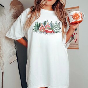 May include: A white short-sleeve t-shirt featuring a graphic print of a red car carrying a Christmas tree in front of red houses and green pine trees. The shirt is paired with a white mug featuring an orange sun graphic with rays.