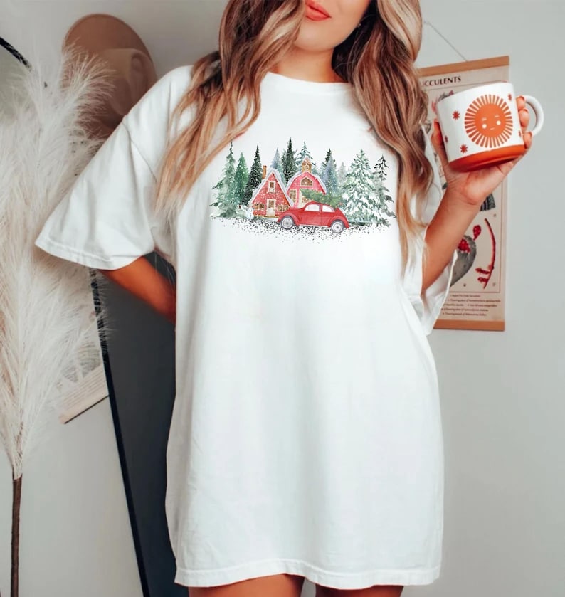 May include: A white short-sleeve t-shirt featuring a graphic print of a red car carrying a Christmas tree in front of red houses and green pine trees. The shirt is paired with a white mug featuring an orange sun graphic with rays.