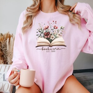 May include: Pale pink sweatshirt with a floral design above an open book. The text "bookworm EST. 1989" is below the book. The sweatshirt is made of a soft, comfortable material.