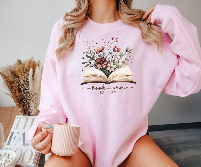May include: Pale pink sweatshirt with a floral design above an open book. The text "bookworm EST. 1989" is below the book. The sweatshirt is made of a soft, comfortable material.