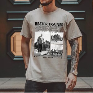 May include: Beige t-shirt with the text "BESTER TRAINER" repeated three times in black. The shirt features a collage of black and white photos. The shirt is a crew neck and has short sleeves.