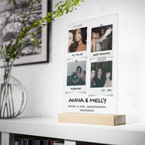 May include: A clear acrylic photo display with four instant photos attached. The photos feature groups of women. The display has a wooden base and text that reads "ANNA & MELLY" and "PARTNER IN CRIME, GEHEIMNISH&Uuml;TERIN, HERZENSMENSCH."