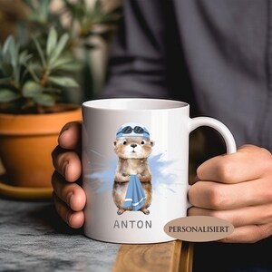 Pode incluir: Caneca de cer&acirc;mica branca com uma lontra de desenho animado usando touca e &oacute;culos de nata&ccedil;&atilde;o, segurando uma toalha azul. O nome "ANTON" &eacute; impresso abaixo da lontra. Um respingo de aquarela azul est&aacute; atr&aacute;s da lontra.