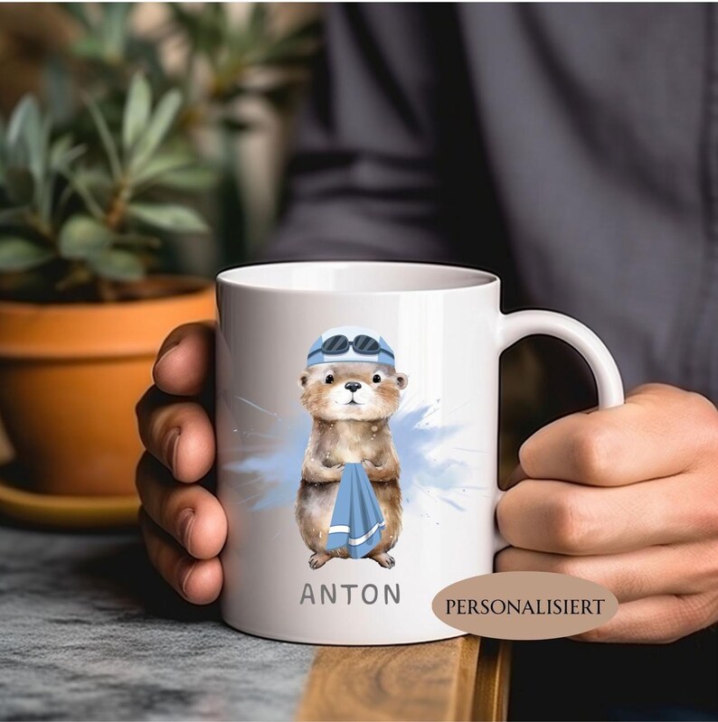 Pode incluir: Caneca de cer&acirc;mica branca com uma lontra de desenho animado usando touca e &oacute;culos de nata&ccedil;&atilde;o, segurando uma toalha azul. O nome "ANTON" &eacute; impresso abaixo da lontra. Um respingo de aquarela azul est&aacute; atr&aacute;s da lontra.