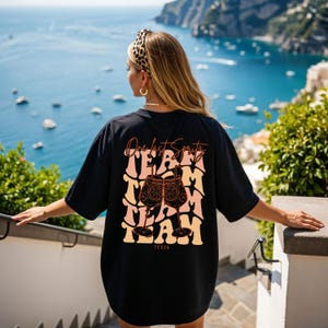 May include: Black oversized t-shirt with the text "One last Spritz" and "TEAM" in a retro font. The graphic includes two cocktail glasses with orange slices. The shirt also has the name "Tessa" at the bottom.