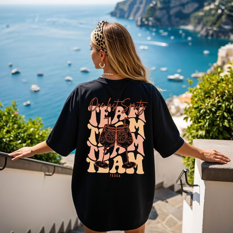May include: Black oversized t-shirt with the text "One last Spritz" and "TEAM" in a retro font. The graphic includes two cocktail glasses with orange slices. The shirt also has the name "Tessa" at the bottom.