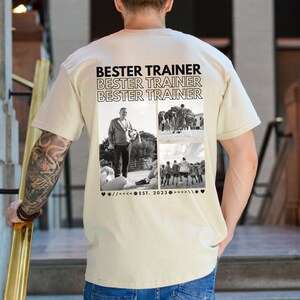 May include: A beige t-shirt featuring the words "BESTER TRAINER" repeated three times. Below the text are three black and white photographs. The bottom of the shirt has the text "EST. 2023" with decorative symbols.