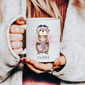 Pode incluir: Caneca de cer&acirc;mica branca com uma ilustra&ccedil;&atilde;o em aquarela de uma lontra usando &oacute;culos de nata&ccedil;&atilde;o e segurando uma toalha. O nome "CLARA" est&aacute; impresso em preto abaixo. A caneca tem um fundo rosa com respingos de aquarela.