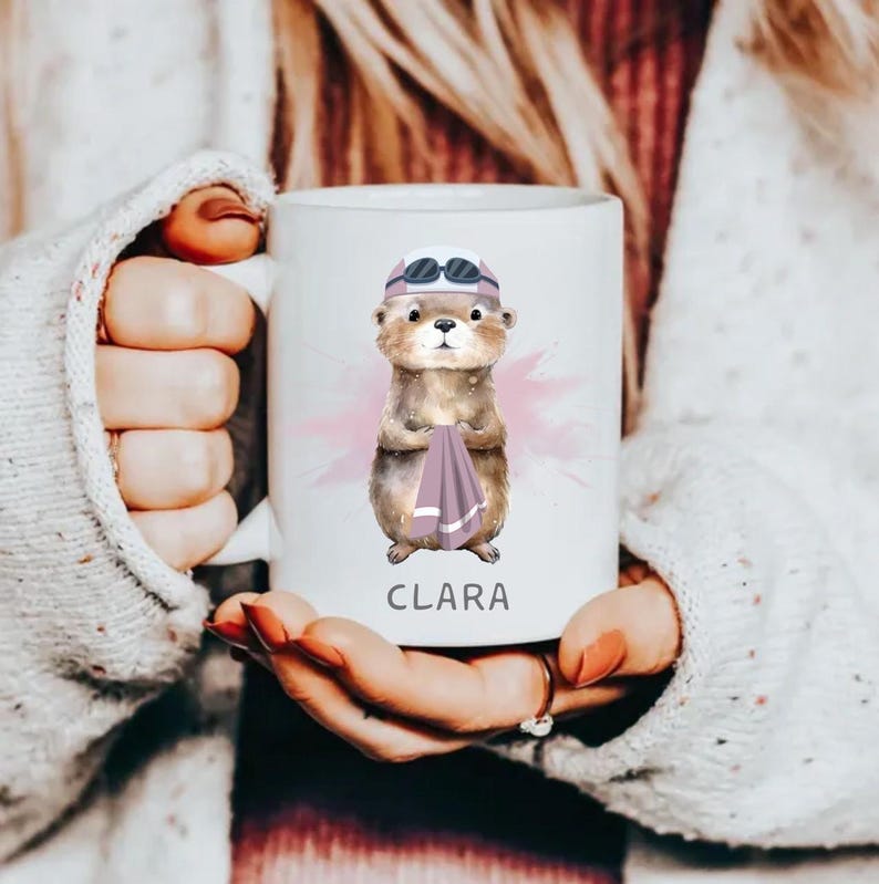 Pode incluir: Caneca de cer&acirc;mica branca com uma ilustra&ccedil;&atilde;o em aquarela de uma lontra usando &oacute;culos de nata&ccedil;&atilde;o e segurando uma toalha. O nome "CLARA" est&aacute; impresso em preto abaixo. A caneca tem um fundo rosa com respingos de aquarela.