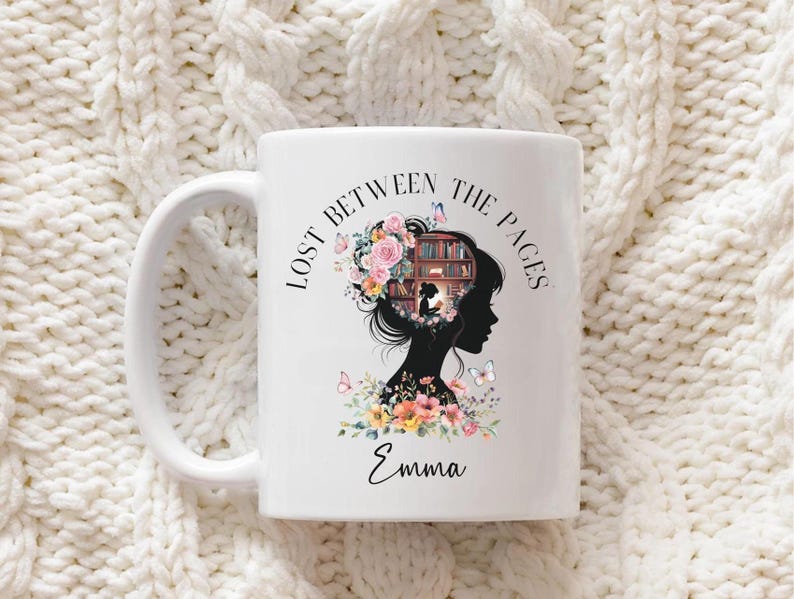 Może przedstawiać: Biały ceramiczny kubek z napisem "LOST BETWEEN THE PAGES" i imieniem "Emma". Kubek przedstawia sylwetkę kobiety z kwiatowym wzorem i p&oacute;łką na książki w jej głowie, z motylami.