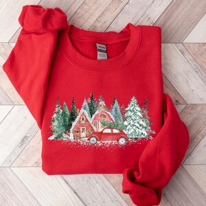 May include: A bright red crewneck sweatshirt featuring a graphic print of a red vintage car parked in front of two red houses surrounded by snow-covered evergreen trees. The design includes a dusting of white snow at the base of the scene.