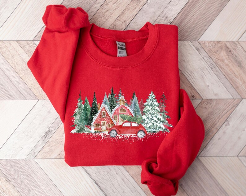 May include: A bright red crewneck sweatshirt featuring a graphic print of a red vintage car parked in front of two red houses surrounded by snow-covered evergreen trees. The design includes a dusting of white snow at the base of the scene.