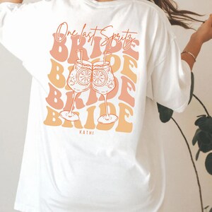 May include: White t-shirt with a back graphic featuring the text "One last Spritz" above the word "Bride" repeated in a stacked design. Two cocktail glasses with orange slices are in the center. The name "Kathi" is at the bottom.