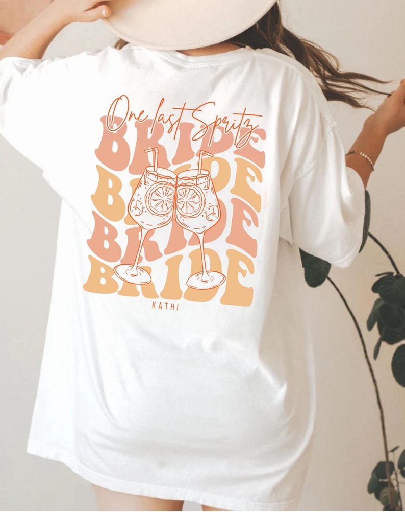 May include: White t-shirt with a back graphic featuring the text "One last Spritz" above the word "Bride" repeated in a stacked design. Two cocktail glasses with orange slices are in the center. The name "Kathi" is at the bottom.