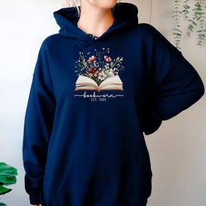 May include: Dark blue hoodie with a floral design above an open book. The cursive text reads "bookworm", with "EST. 1989" underneath. The hoodie has a front pocket and a drawstring hood.