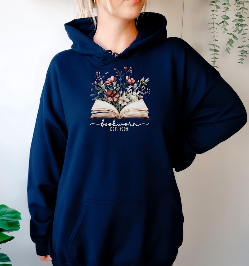 May include: Dark blue hoodie with a floral design above an open book. The cursive text reads "bookworm", with "EST. 1989" underneath. The hoodie has a front pocket and a drawstring hood.