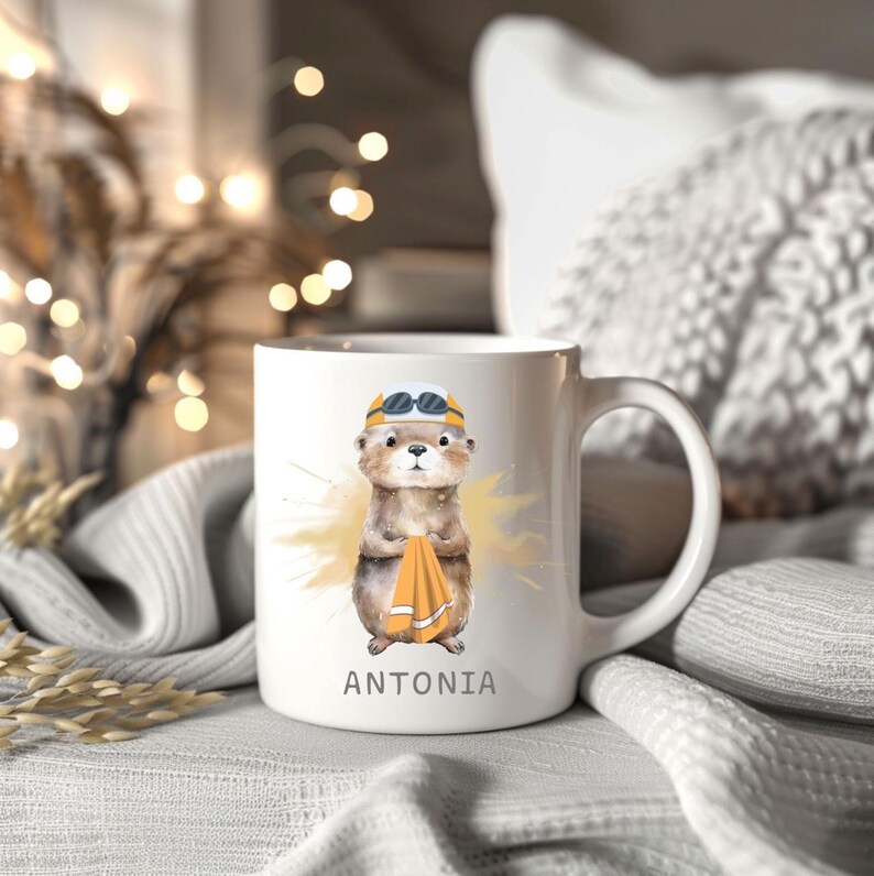 Pode incluir: Caneca de cer&acirc;mica branca com uma lontra de desenho animado usando &oacute;culos de nata&ccedil;&atilde;o e uma touca de banho amarela, segurando uma toalha laranja. O nome "ANTONIA" est&aacute; impresso abaixo. A caneca tem uma al&ccedil;a curva.