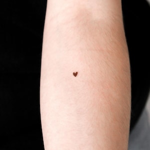 May include: A tiny, dark brown heart tattoo on the inner arm. The tattoo is a simple, solid shape, and the skin around it is light-toned. The background is a dark, blurred area.