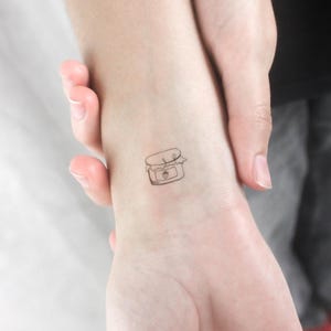 Può includere: Un semplice tatuaggio a linea di un barattolo di marmellata con una fragola sull'etichetta, sul polso di una persona.
