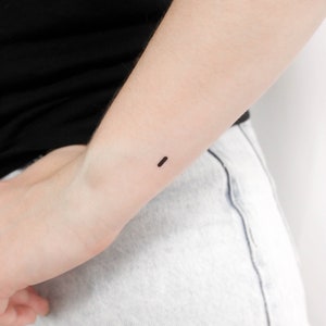May include: A close-up of a person's wrist with a small, simple black line tattoo.