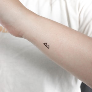 Puede incluir: Un simple tatuaje de línea negra de dos triángulos en la muñeca de una persona.