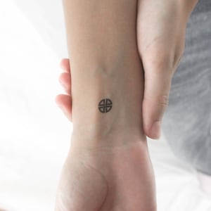 May include: A small, black tattoo of a four-part circular design on a wrist. The tattoo is centered on the wrist and appears to be freshly applied. The skin around the tattoo is light-toned.