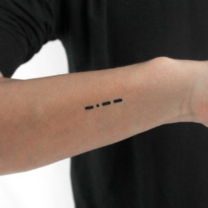 May include: A close-up of a person's forearm with a simple black tattoo of three dots and a dash. The tattoo is a Morse code representation of the letter 'S'.