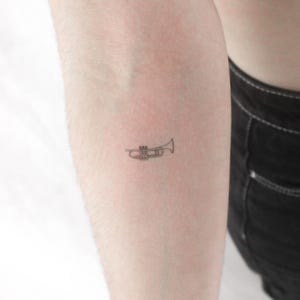 Puede incluir: Un pequeño tatuaje minimalista de una trompeta. El diseño es una ilustración de líneas finas del instrumento de viento, centrándose en sus características esenciales. El tatuaje está en el brazo.