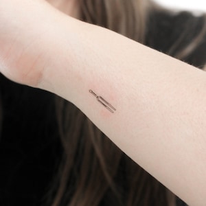 May include: A small, simple line drawing tattoo of a tuning fork on a person's wrist.
