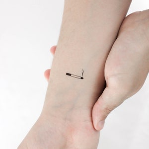 May include: A simple black and white tattoo design of a cigarette with smoke on a person's wrist.