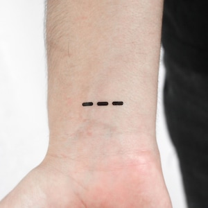 May include: A close-up of a wrist with a small, minimalist tattoo. The tattoo consists of three evenly spaced, horizontal black lines. The skin tone is light, and the tattoo is centered on the wrist.