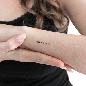 May include: A simple black line tattoo on a person's wrist. The tattoo is a short line followed by three dots.