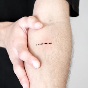 May include: A black temporary tattoo of Morse code dots and dashes on a person's forearm. The tattoo is a simple, minimalist design, with the dots and dashes evenly spaced. The skin is light-toned.