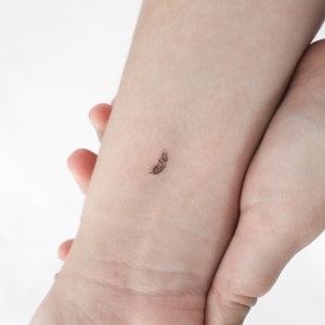 May include: A small, simple black feather tattoo on a person's wrist.