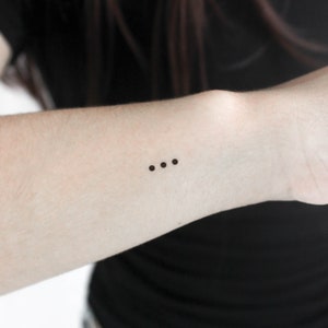 May include: A close-up of a person's wrist with a small tattoo of three black dots.